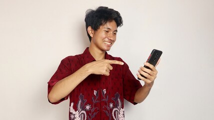 happy young handsome Asian man in batik shirt posing pointing at smartphone