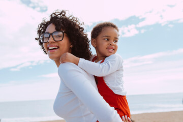 Smiling beautiful mother and her daugther enjoy together piggyback on beach, Love, vacation, happy family, ocean and tourism concept. Child hugs woman outdoor, mockup space. - Powered by Adobe