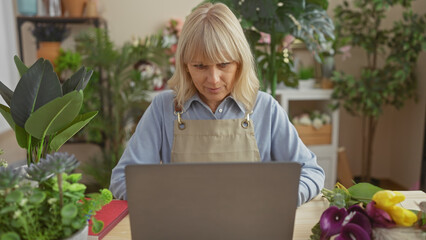 A focused blonde woman using a laptop amid vibrant flowers in a florist's shop