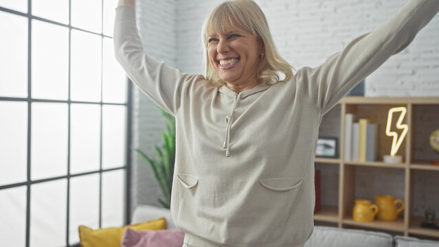 A joyful mature woman celebrates indoors, raising her arms in a modern living room setup.