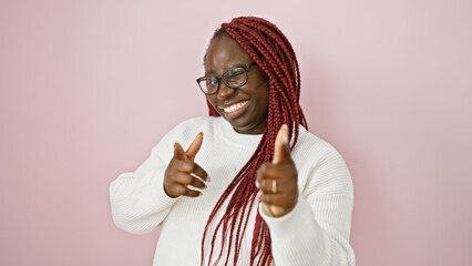 Smiling african woman with braids winking and pointing over isolated pink wall background
