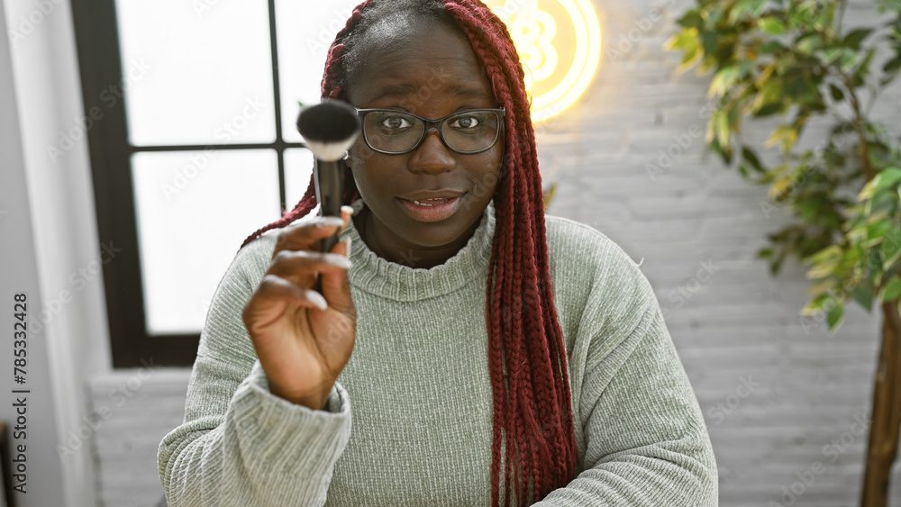 Sticker a beautiful african american woman with braids wearing glasses holds a makeup brush in a cozy indoor