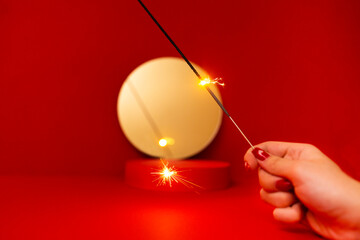 A lit sparkler in the hand of a young woman with a red manicure. Red podium and round mirror on the background. - Powered by Adobe