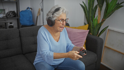 Elderly woman with headphones playing video games in a cozy living room, showcasing active senior...
