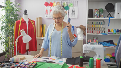 A senior woman focuses on a phone call, surrounded by colorful fabrics and sewing equipment in a...