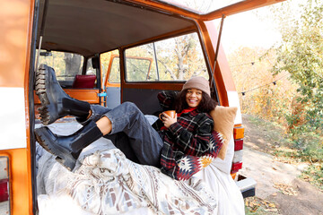 Young african american woman sitting at table near car in autumn forest and drinking hot tea, dreamy mixed-race female relaxing around campsite, spending time in nature. Camping, travel and vanlife - Powered by Adobe