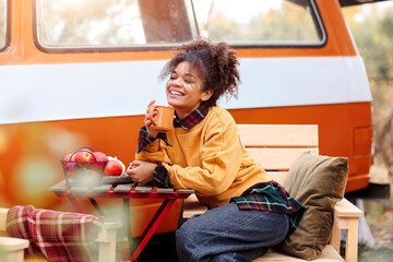 Young african american woman sitting at table near car in autumn forest and drinking hot tea, dreamy mixed-race female relaxing around campsite, spending time in nature. Camping, travel and vanlife - Powered by Adobe