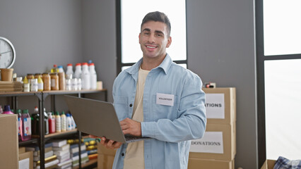 Young, confident hispanic man volunteers with a smile, using laptop at a buzzing charity center,...