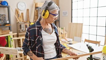 Serious, grey-haired middle age woman carpenter deeply invested in her woodwork, fervently looking...