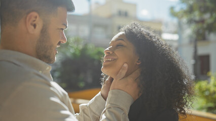 A man tenderly touches the face of a smiling woman in a sunlit outdoor urban park.