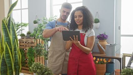Man and woman in aprons reviewing tablet inside plant-packed flower shop, portraying teamwork and retail business.