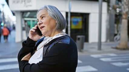 Senior woman conversing on mobile phone while standing on a busy city sidewalk. - obrazy, fototapety, plakaty