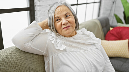 A thoughtful senior woman relaxes at home in a comfortable living room setting, exuding a sense of...
