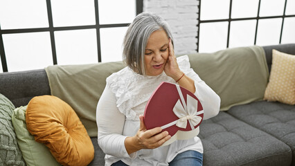 Senior woman surprised by heart-shaped gift box while sitting on a sofa in a cozy living room.