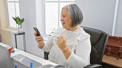 Joyful mature woman celebrating success with smartphone at office desk.