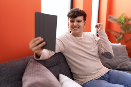 Young non binary man using touchpad sitting on the sofa screaming proud, celebrating victory and success very excited with raised arm