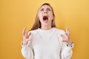Young caucasian woman wearing white sweater over yellow background crazy and mad shouting and...