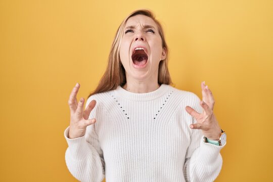 Young caucasian woman wearing white sweater over yellow background crazy and mad shouting and yelling with aggressive expression and arms raised. frustration concept.