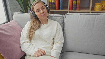 A relaxed young woman enjoys music on headphones while sitting on a sofa in a cozy living room.