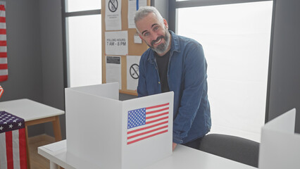 A smiling bearded man voting in an american polling station beside usa flags.