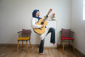 Asian young girl in hijab emotionally playing the acoustic guitar standing with one leg raised.