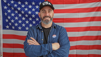 Smiling bearded man in denim with 'voted' sticker, crossed arms, american flag background,...