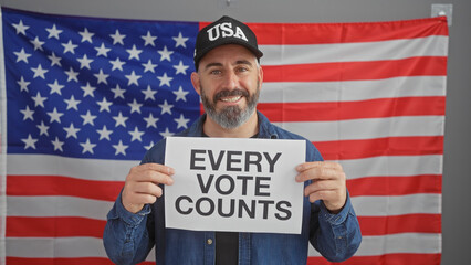 Smiling bearded man holding a 'every vote counts' sign with american flag backdrop in an office...