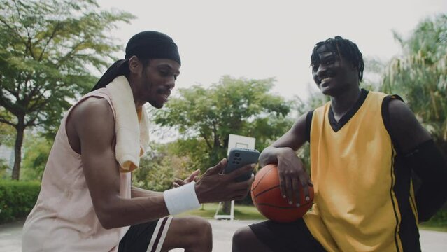 Handheld shot of two black male sporty friends checking smartphone after basketball training on outdoor playground