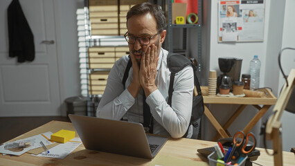 Mature man with beard in office looks distressed contemplating evidence on cluttered desk.
