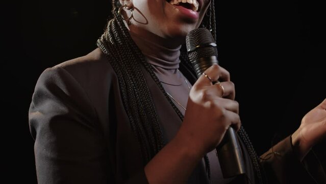 Chest up of young Black woman with braided hairstyle standing with microphone on dark stage and speaking while performing standup comedy monologue