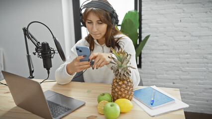 Young woman with headphones in a radio studio looking at phone by microphone and laptop, evoking a...