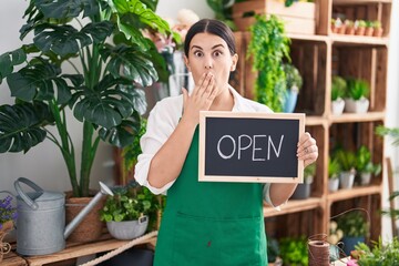 Young hispanic woman working at florist holding open sign covering mouth with hand, shocked and...