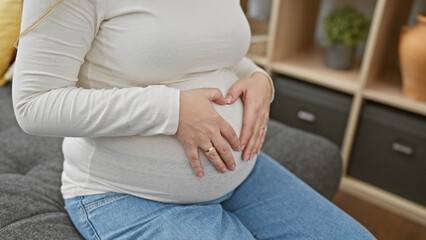 A young hispanic pregnant woman makes a heart gesture over her belly in an indoor setting.