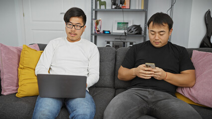 Two asian men relax on a sofa, one using a laptop and the other a smartphone, in a modern living room.