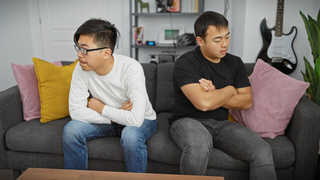 Two asian men sitting apart on a grey couch in a modern living room, seeming distant and pensive.