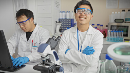 Two men working together in a laboratory setting, one using a microscope and the other typing on a computer.