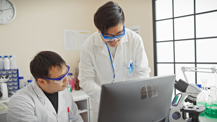 Two men in lab coats work together in a bright laboratory setting, examining data on a computer screen.