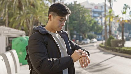 A young asian man checks the time on his wristwatch while walking on a sunny urban street.