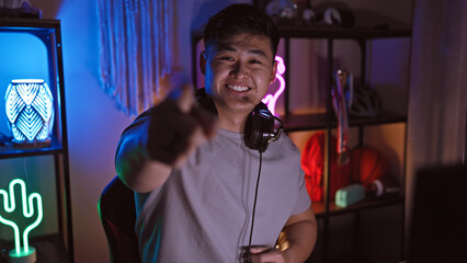 A happy young asian man in a gaming room pointing at the camera with led lights at night.