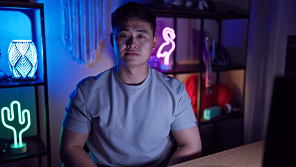 Handsome asian man with neutral expression in a dark illuminated gaming room at home.