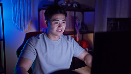 A smiling asian man in a dark gaming room lit by colorful lights, enjoying his leisure time at home...