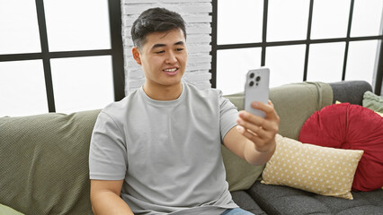 A young asian man takes a selfie with his smartphone in a cozy, stylish living room