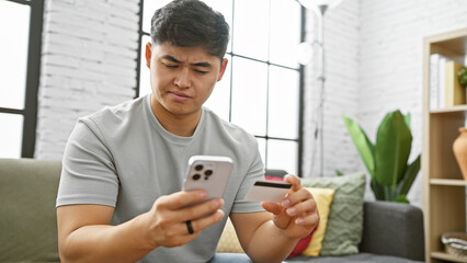 A young asian man analyzes his credit card while using a smartphone in a modern living room.