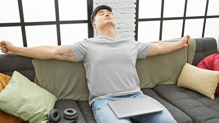 A relaxed asian man stretching on a sofa in a modern living room with headphones and a laptop.