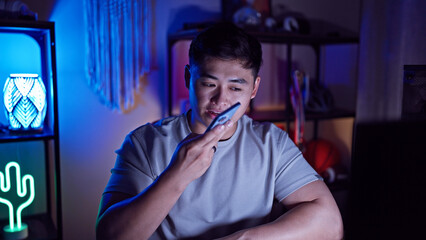 A young asian man sending a voice message indoors at night in a room with neon lighting.
