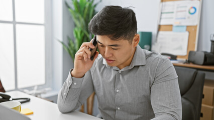 A focused young asian man in an office setting talks on the phone, suggesting business, communication, or customer service.