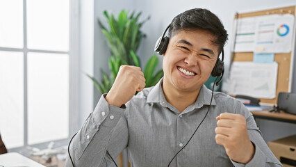 A cheerful asian man wearing a headset celebrating success in an office environment.