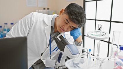 A young asian man in a lab coat appears exhausted while working with a microscope in a laboratory setting.
