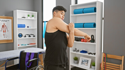 An asian man stretches his arm in a modern rehabilitation clinic's gym, indicating a healthcare or physical therapy setting.