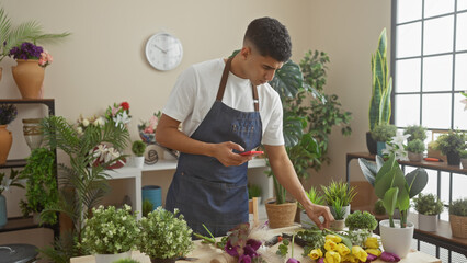 A young man uses a smartphone while working among colorful flowers in an indoor flower shop.